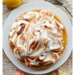 A homemade Lemon Meringue Tart with a buttery pastry crust filled with silky fresh lemon curd and topped with toasted Italian meringue, served on a white cake stand with fresh lemons and soft summer décor.