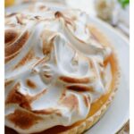 A homemade Lemon Meringue Tart with a buttery pastry crust filled with silky fresh lemon curd and topped with toasted Italian meringue, served on a white cake stand with fresh lemons and soft summer décor.
