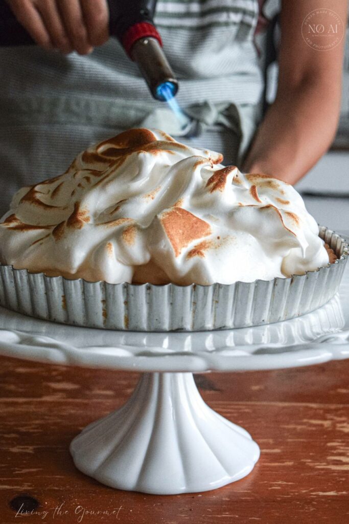 A homemade Lemon Meringue Tart with a buttery pastry crust filled with silky fresh lemon curd and topped with toasted Italian meringue, served on a white cake stand with fresh lemons and soft summer décor.