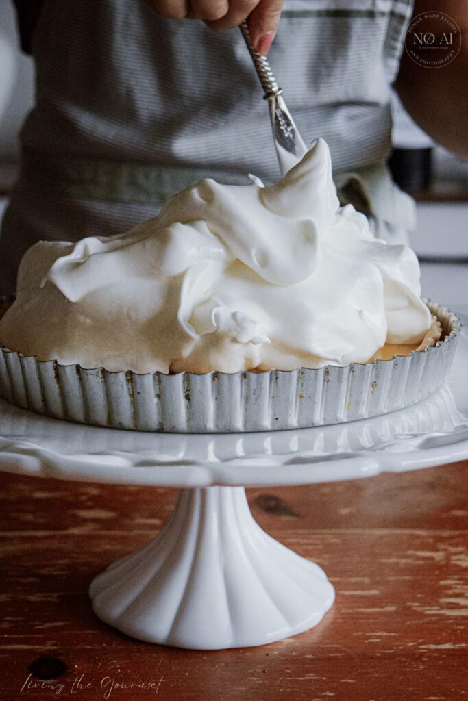 A homemade Lemon Meringue Tart with a buttery pastry crust filled with silky fresh lemon curd and topped with toasted Italian meringue, served on a white cake stand with fresh lemons and soft summer décor.