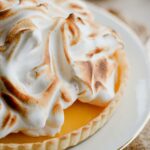 A homemade Lemon Meringue Tart with a buttery pastry crust filled with silky fresh lemon curd and topped with toasted Italian meringue, served on a white cake stand with fresh lemons and soft summer décor.