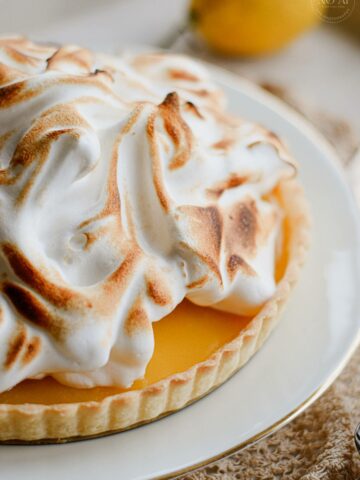 A homemade Lemon Meringue Tart with a buttery pastry crust filled with silky fresh lemon curd and topped with toasted Italian meringue, served on a white cake stand with fresh lemons and soft summer décor.